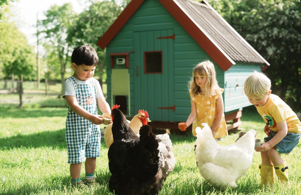 Three young children smiling and feeding a group of chickens in front of a decorated chicken coop